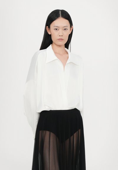 Blusa blanca de manga larga con collar, combinada con una falda negra transparente con pliegues y cintura alta. Diseño minimalista con texturas suaves.