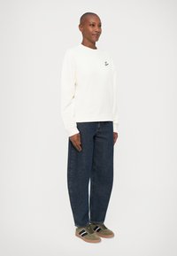Felpa bianca con collo a giro, dotata di un piccolo logo nero, abbinata a jeans blu scuro dal taglio ampio e sneakers in camoscio verde con strisce nere.
