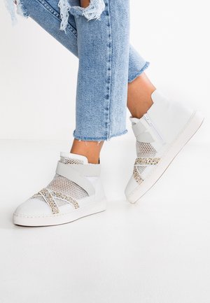 Fødder iført hvide høje sneakers med mesh-paneler og glitrende stropper, kombineret med lyseblå revnede jeans med frynset kant.