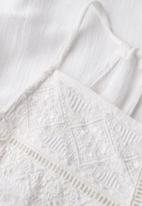 Witte stof met ingewikkelde borduursels met diamanten en geometrische patronen, aangevuld met gestructureerde, geweven details en dunne bandjes.