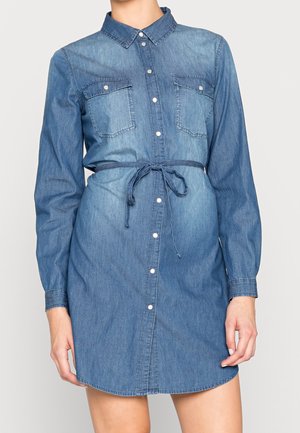 Femme portant une robe-chemise en jean bleu à boutons, manches longues, deux poches poitrine et une ceinture nouée à la taille.