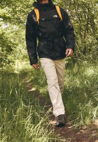Chaqueta negra impermeable con correas naranjas, pantalones blancos y botas de senderismo; caminando por un sendero de tierra rodeado de hierba verde y árboles.