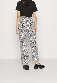 Pantalones de pierna ancha en blanco y negro con un diseño abstracto de olas. Presentan una cinturilla alta con la marca "RAGGED". Combinados con zapatos negros.