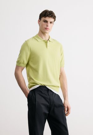 Νέος άνδρας με ανοιχτοπράσινο πόλο μπλουζάκι με κοντά μανίκια, φορεμένο πάνω από λευκό εσώρουχο και μαύρο παντελόνι, στέκεται μπροστά σε απλό λευκό φόντο.