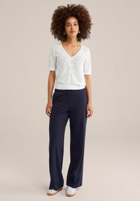 Cardigan blanc en dentelle à manches courtes avec un col en V et une fermeture à boutons, associé à un pantalon large bleu marine et des baskets blanches. Couleur unie, texture lisse.