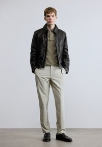 Μαύρο δερμάτινο μπουφάν, χακί πόλο πουκάμισο, καλοραμμένο κρεμ παντελόνι και μαύρα παπούτσια. Το μοντέλο στέκεται ίσια μπροστά σε απλό φόντο.
