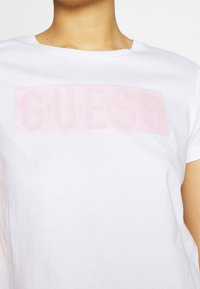 Bílá bavlněná tričko s velkým, lesklým růžovým logem "GUESS" potisknutým přes hrudník, krátké rukávy a kulatý výstřih.