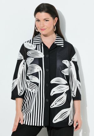 Kobieta ubrana w czarną koszulę zapinaną na guziki z białymi abstrakcyjnymi wzorami liści i pasków, stojąca na tle jednolitego jasnego tła.