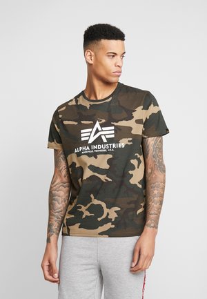 T-shirt à motif camouflage dans des teintes de vert et de marron avec un logo blanc portant "ALPHA INDUSTRIES" sur le devant. Manches courtes, col rond.