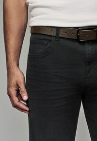 Vaqueros de mezclilla negra con un corte ajustado, costuras detalladas, bolsillos frontales y un cinturón de cuero marrón con agujeros visibles y una hebilla de metal.