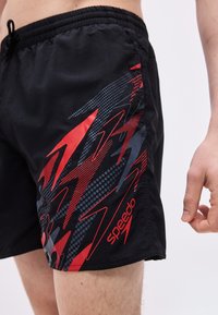 Μαύρα μαγιό βερμούδα με κόκκινα και γκρι αφηρημένα σχέδια και το λογότυπο "Speedo", φορεμένα από έναν άνδρα με ορατά γυμνά πόδια και χέρι.