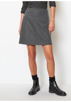 Kvinde iført en knælængde grå nederdel, sorte ankelstøvler, sorte sokker og en langærmet skjorte med striber, stående foran en hvid baggrund.