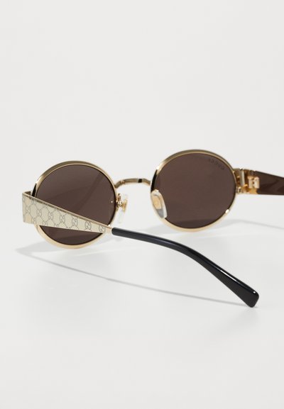 Gafas de sol redondas con lentes marrones, montura de metal dorado y brazos detallados con un patrón beige que presenta el logo de la marca. Puntas de las patas negras.