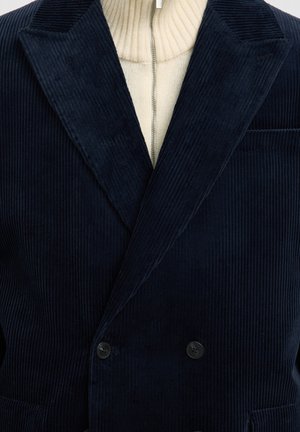 Navy corduroy dubbele-breasted blazer met brede revers, gestructureerde stof en twee zwarte knopen. Wit geribde coltrui zichtbaar eronder.