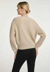 Kobieta z blond włosami w niskim koku, ubrana w beżowy sweter z dzianiny i czarne spodnie, stojąca z jedną ręką na biodrze, zwrócona bokiem.