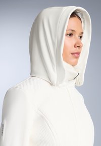 Jachetă albă cu glugă, cu o textură netedă, având un model geometric pe piept și o căptușeală moale în jurul gulerului.