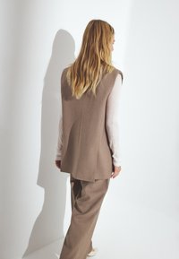 Beige sleeveless vest layered over a light long-sleeve top, paired with matching loose trousers. Smooth fabric with a tailored cut.