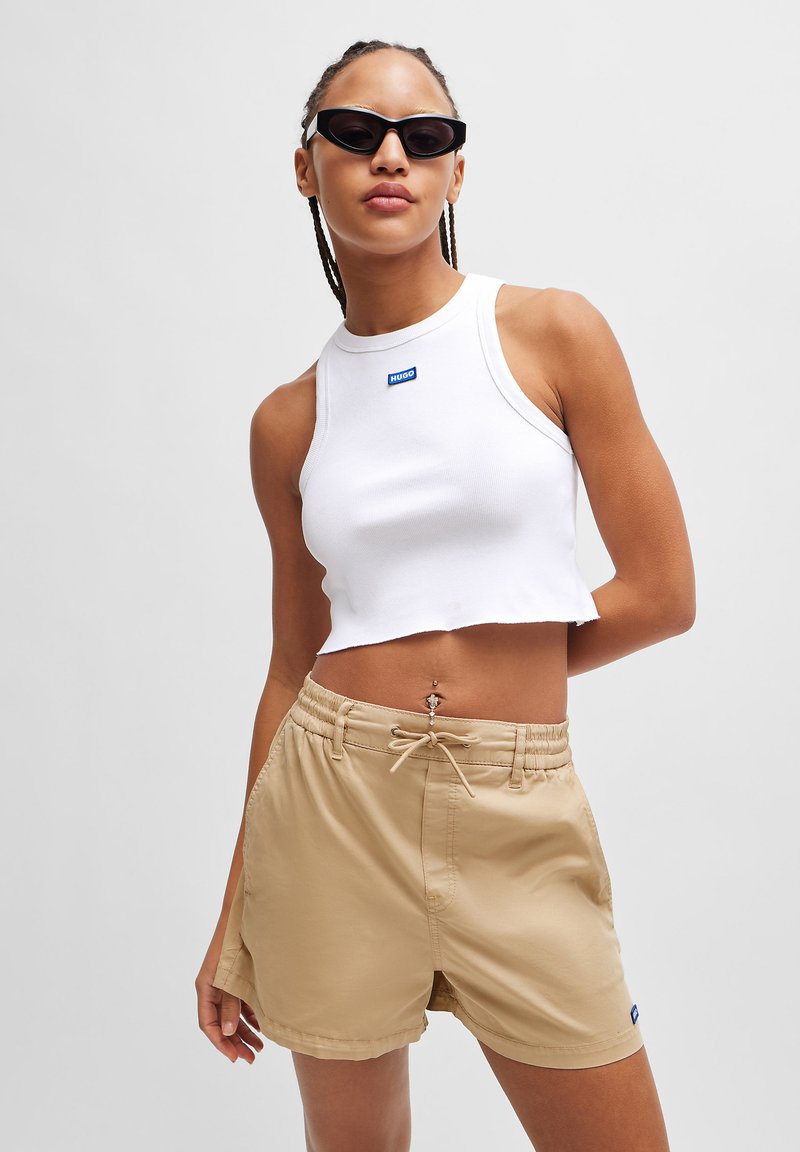 Witte geribde cropped tanktop met blauw logo, gecombineerd met beige shorts met een elastische tailleband en een koord. Zwarte zonnebril.