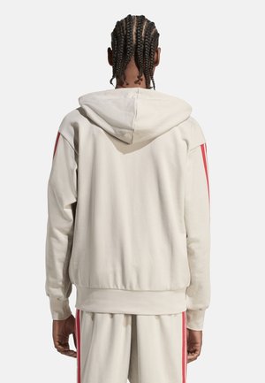 Man met gevlochten haar draagt een off-white hoodie en bijpassende broek met rode strepen, kijkt weg op een effen achtergrond.