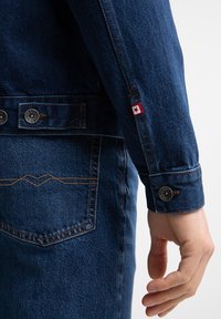 Denim jakna s temno modrim blagom, kontrastnim šivanjem, kovinskimi gumbi in majhno rdečo oznako z zvezdo na rokavu.