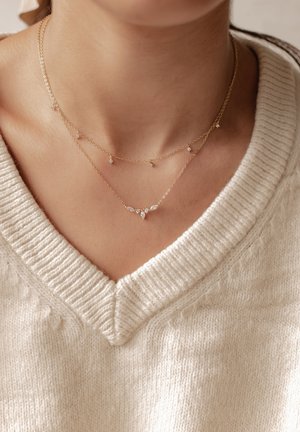 Goldfarbene Schichtenketten mit kleinen, diamantähnlichen Akzenten, unterschiedlicher Längen und zarten Kettenmustern, die zu einem leichten Pullover passen.