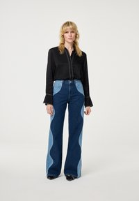 Zwarte blouse met lange mouwen en een knopsluiting aan de voorkant, gecombineerd met hooggetailleerde blauwe jeans met golvende lichtblauwe zijkanten.