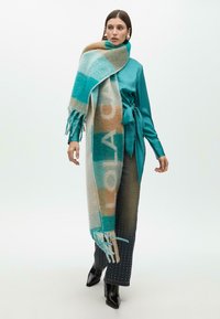 Šála z vlněné směsi v tyrkysově modré, béžové a modré kostkované vzoru. Má třásně. Modelka má na sobě saténovou blůzu v barvě odlady a tečkované džíny.