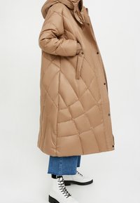 Tan gewatteerde lange jas met een capuchon, met zijzakken en een drukknoopsluiting. Draagt over blauwe jeans en witte veterschoenen.