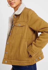 Musztardowożółta kurtka z kołnierzem z baranka, metalowymi guzikami, bocznymi kieszeniami i przemyślaną konstrukcją. Materiał bawełniany o fakturowanym wykończeniu.