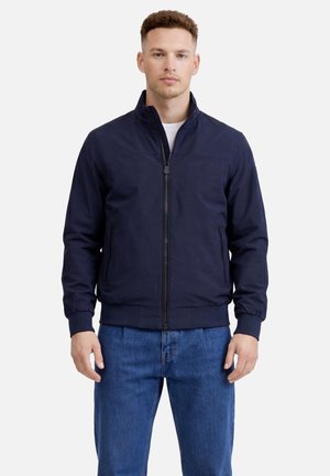 Uomo con giacca blu navy con zip e jeans blu, in piedi con le braccia rilassate davanti a uno sfondo chiaro e semplice.
