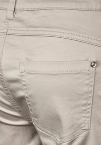 Beige bomullbyxor med en bakficka med en blank metallknapp, sömlös sömnad och en slät, något strukturert yta.