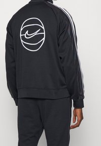 Chaqueta de chándal negra de Nike con logo blanco, textura lisa y acentos a rayas en las mangas. Cuenta con bolsillos con cremallera y puños de canalé.