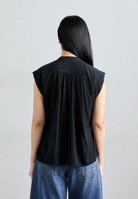 Blouse noire à manches courtes en tissu léger, avec un design à dos plissé. Portée avec un jean en denim bleu taille haute.