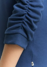 Blauer Strickpullover mit gerafften ÄrmelDetails und gerippten Bündchen. Enthält ein kleines weißes Etikett an der Naht. Weiche Textur, figurbetontes Design.