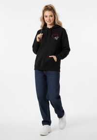 Felpa nera con cappuccio, decorata con una grafica di un cuore e mani, con tasca frontale. Abbinata a jeans blu scuro e sneakers bianche.