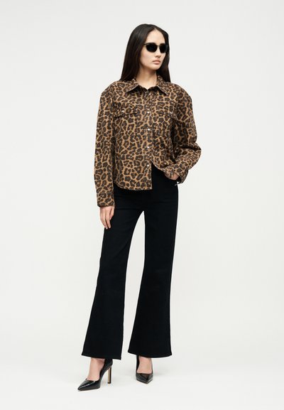 Woman wearing black sunglasses, leopard print button-up shirt, black flared pants, and black high heels, standing with one hand in pocket.