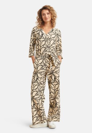 Beige jumpsuit met een zwart bloemenpatroon; heeft een V-hals, driekwart mouwen, een strikceintuur en een wijde broekspijp. Het textiel lijkt glad.