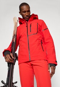 Lys rød ski jakke med vanntett materiale, flere glidelåslommer, justerbar hette og kontrasterende svarte detaljer. Skistav holdt i hånden.