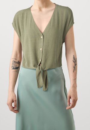 Blouse vert olive à col en V, manches courtes, fermeture à boutons ; nouée à la taille. Associée à une jupe légère en tissu soyeux vert clair.