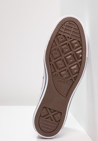 Braune Gummisohle eines Sneakers mit einem geometrischen Profil, weißer Umrandung und Branding in der Nähe der Ferse.