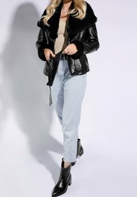 Giacca di pelle nera con collo e polsini in pelliccia sintetica, abbinata a jeans azzurri e stivaletti neri alla caviglia. La giacca ha un design aderente.