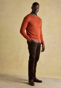 Muž stojaci s rukami v vreckách, oblečený v oranžovom svetri cez károvanú košeľu, hnedých nohaviciach a hnedých loafroch na béžovom pozadí.