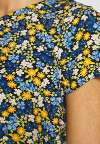 Tessuto a fantasia floreale con sfondo nero, caratterizzato da fiori gialli, blu e bianchi. Texture morbida con maniche corte e una leggera lucentezza.