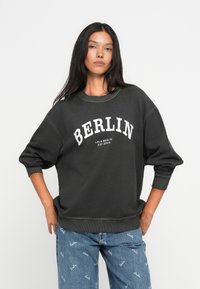 Μαύρη βαμβακερή μπλούζα με εκτύπωση "BERLIN" σε λευκό, με στρογγυλή λαιμόκοψη και χαλαρές μανίκες. Συνδυασμένη με ανοιχτό μπλε τζιν.