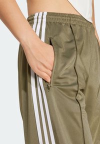 Olivově zelené teplákové kalhoty s bílými pruhy po stranách, s kapsou na zip a hladkým lesklým povrchem. Viditelný elastický pás.
