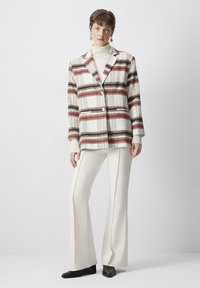 Blazer a righe in crema, marrone e rosso con due bottoni e tasche, abbinato a pantaloni bianchi a zampa d'elefante e stivaletti neri alla caviglia.