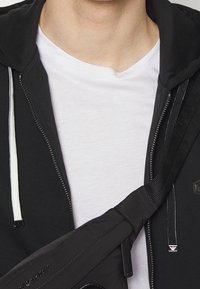 Persona che indossa una maglietta bianca sotto una felpa nera con zip, con lacci bianchi e una tracolla nera del borsello appesa sul petto.