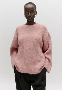 Rosa gestrickter Pullover mit Rundhalsausschnitt, lockerer Passform und strukturiertem Muster. Lange Ärmel und ein weiches, flauschiges Finish. Kombiniert mit einer schwarzen Hose.