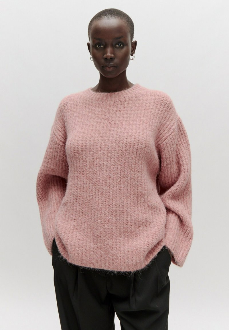 Rosa gestrickter Pullover mit Rundhalsausschnitt, lockerer Passform und strukturiertem Muster. Lange Ärmel und ein weiches, flauschiges Finish. Kombiniert mit einer schwarzen Hose.