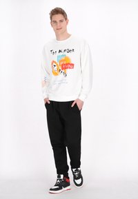 Vit sweatshirt med färggrafisk text, ihop med svarta sweatpants och svarta högskurna sneakers med röda accenter och vita sulor.